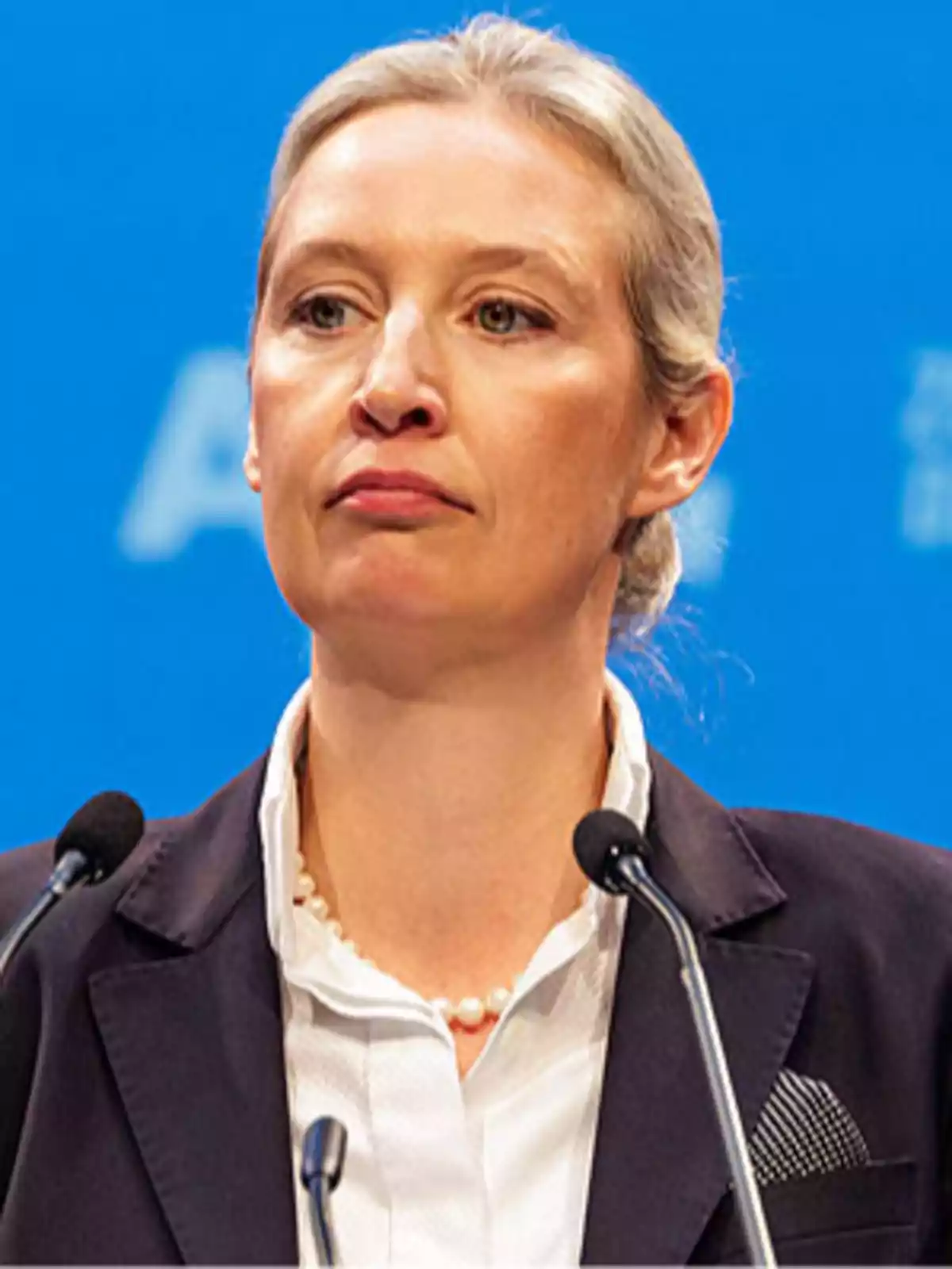 A blonde-haired person in a dark suit speaking in front of a microphone with a blue background.