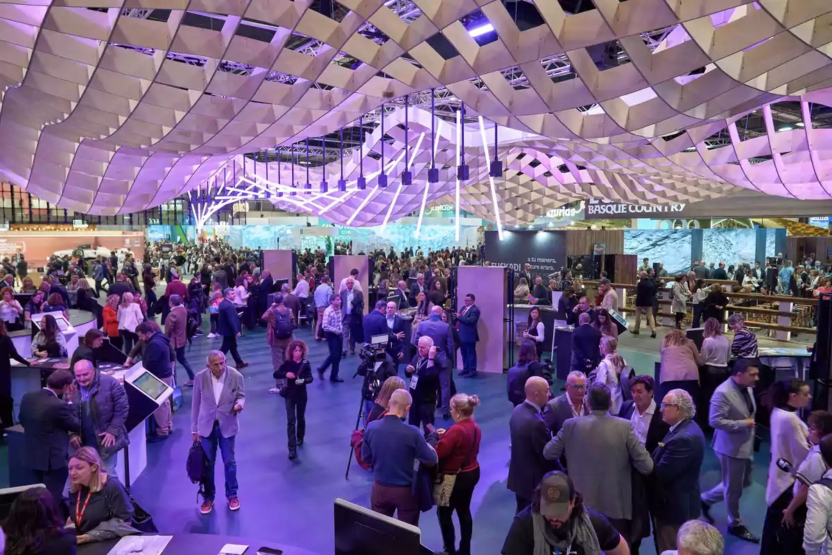 A crowded event with many people interacting under a decorative ceiling lit in purple tones.