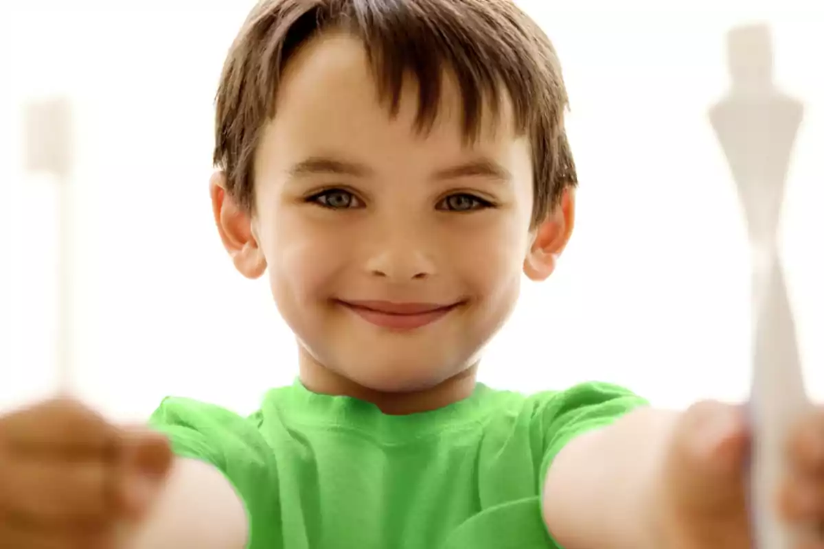 Un niño sonriente con una camiseta verde sosteniendo dos cepillos de dientes.