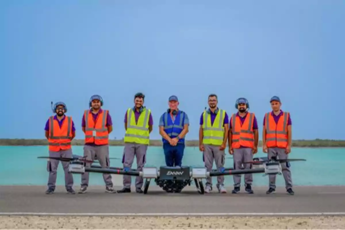 Un grupo de personas con chalecos de seguridad de colores brillantes posando frente a un dron en una pista cerca del agua.