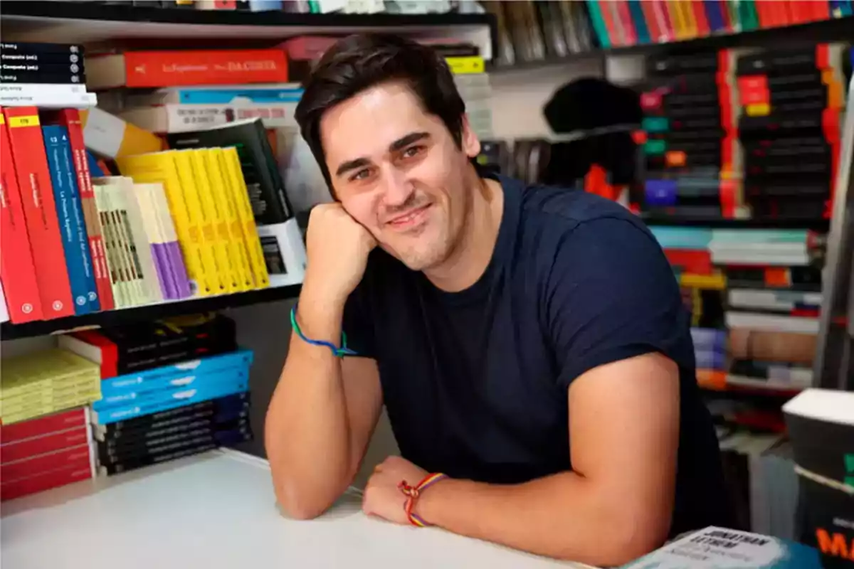 Un hombre sonriente con camiseta azul está apoyado en una mesa rodeado de estantes llenos de libros.