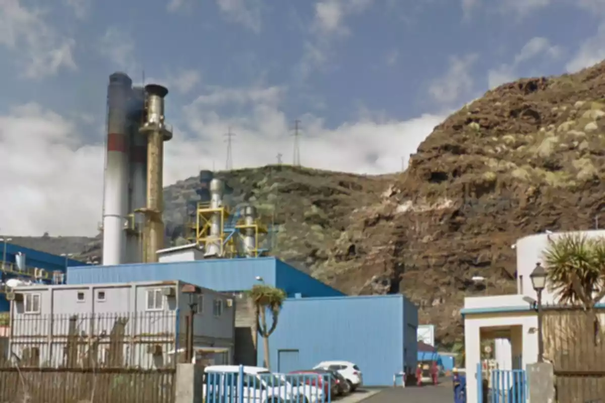 Vista de una planta industrial con edificios azules, chimeneas y colinas rocosas al fondo en un día parcialmente nublado