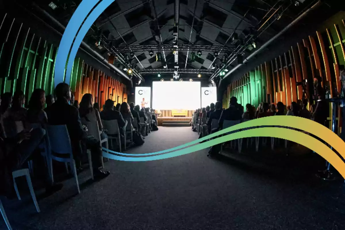 Personas sentadas en una sala de conferencias con iluminación colorida y una pantalla grande al frente.