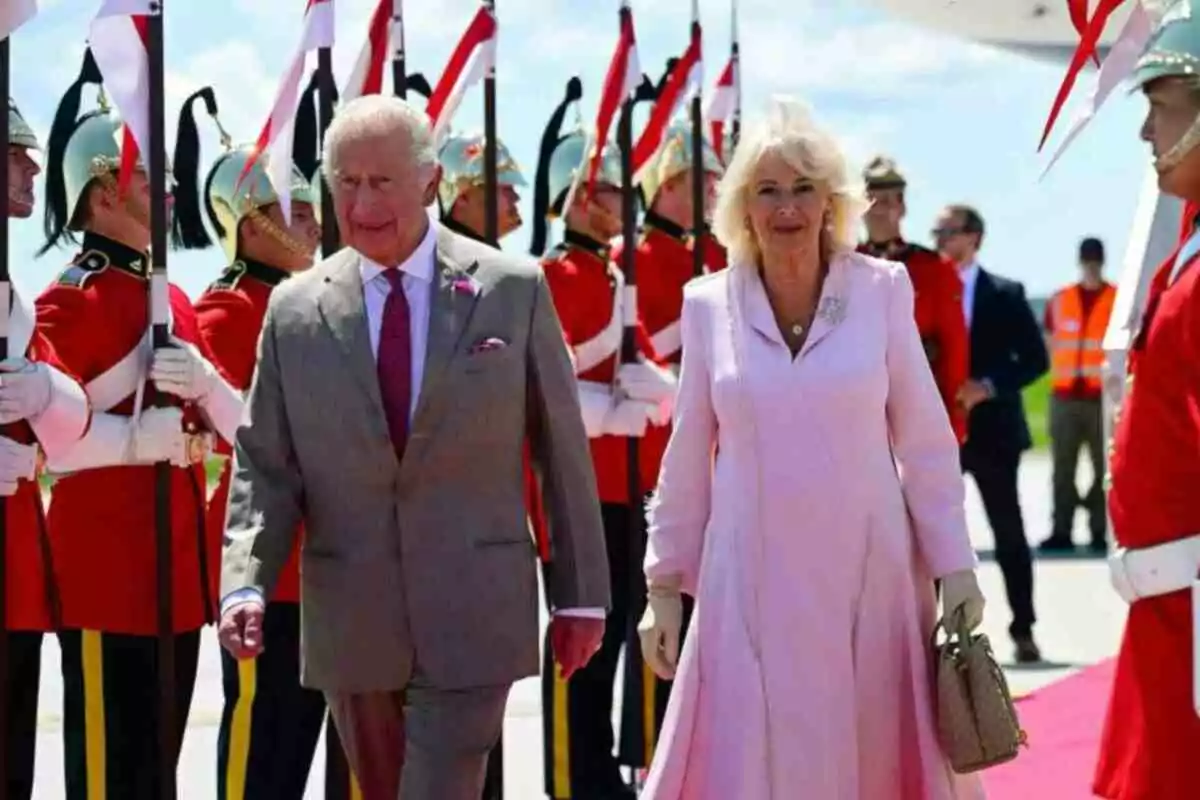 El rey Carlos III y la reina consorte Camila elegantemente vestidos caminan por una alfombra roja mientras soldados con uniformes rojos y cascos plateados forman una guardia de honor a su alrededor El rey Carlos III y la reina consorte Camila elegantemente vestidos caminan por una alfombra roja mientras soldados con uniformes rojos y cascos plateados forman una guardia de honor a su alrededor