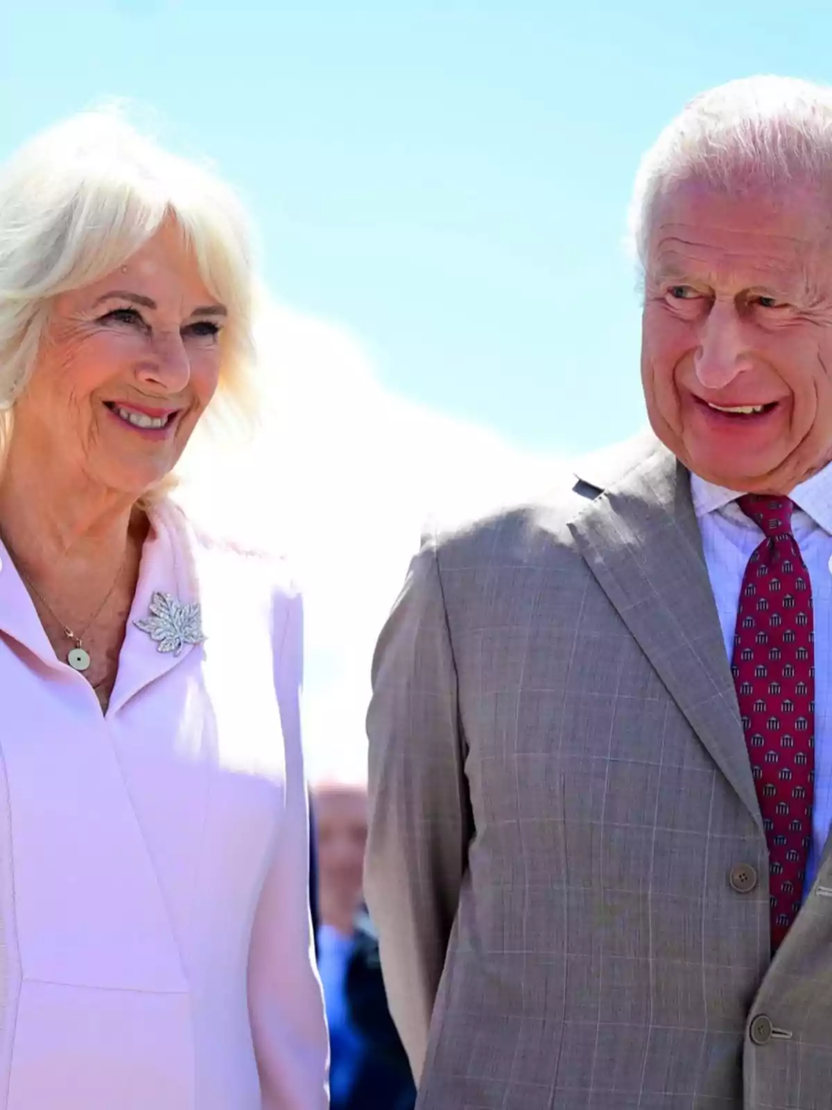 King Charles III and Queen Camila smile while standing outdoors under a blue sky. King Charles III and Queen Camila smile while standing outdoors under a blue sky.