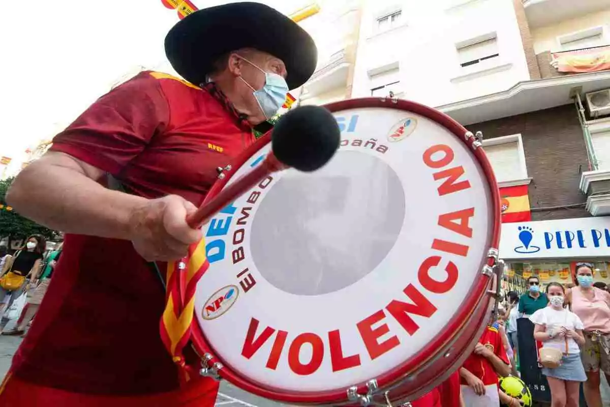 Manolo El del Bombo con sombrero y mascarilla toca un bombo que tiene un mensaje contra la violencia mientras personas observan en la calle. Manolo El del Bombo con sombrero y mascarilla toca un bombo que tiene un mensaje contra la violencia mientras personas observan en la calle.