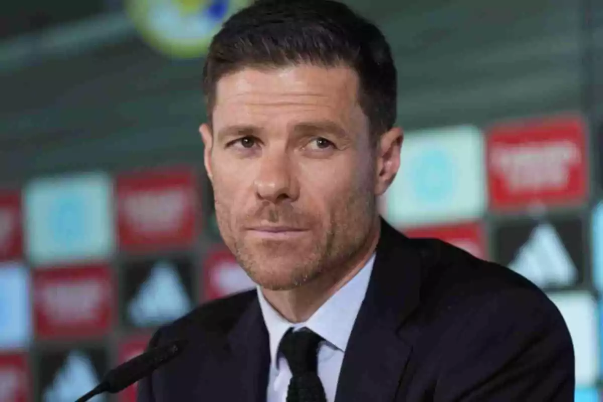 Xabi Alonso con cabello corto y barba recortada usando traje oscuro en una conferencia de prensa con fondo de logotipos desenfocados Xabi Alonso con cabello corto y barba recortada usando traje oscuro en una conferencia de prensa con fondo de logotipos desenfocados