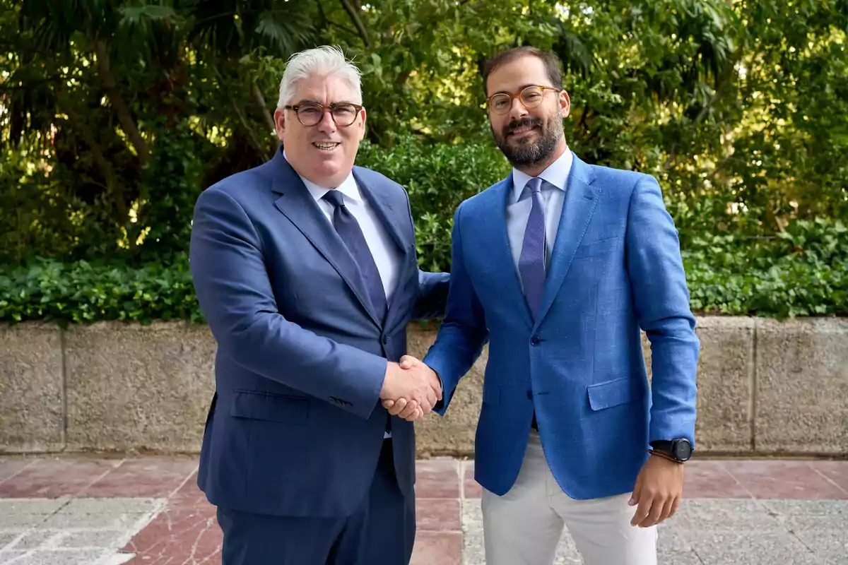 Dos hombres de negocios estrechándose la mano y sonriendo, ambos vestidos con trajes formales, uno de ellos con gafas y cabello canoso, el otro con barba y gafas, en un entorno al aire libre con vegetación de fondo.