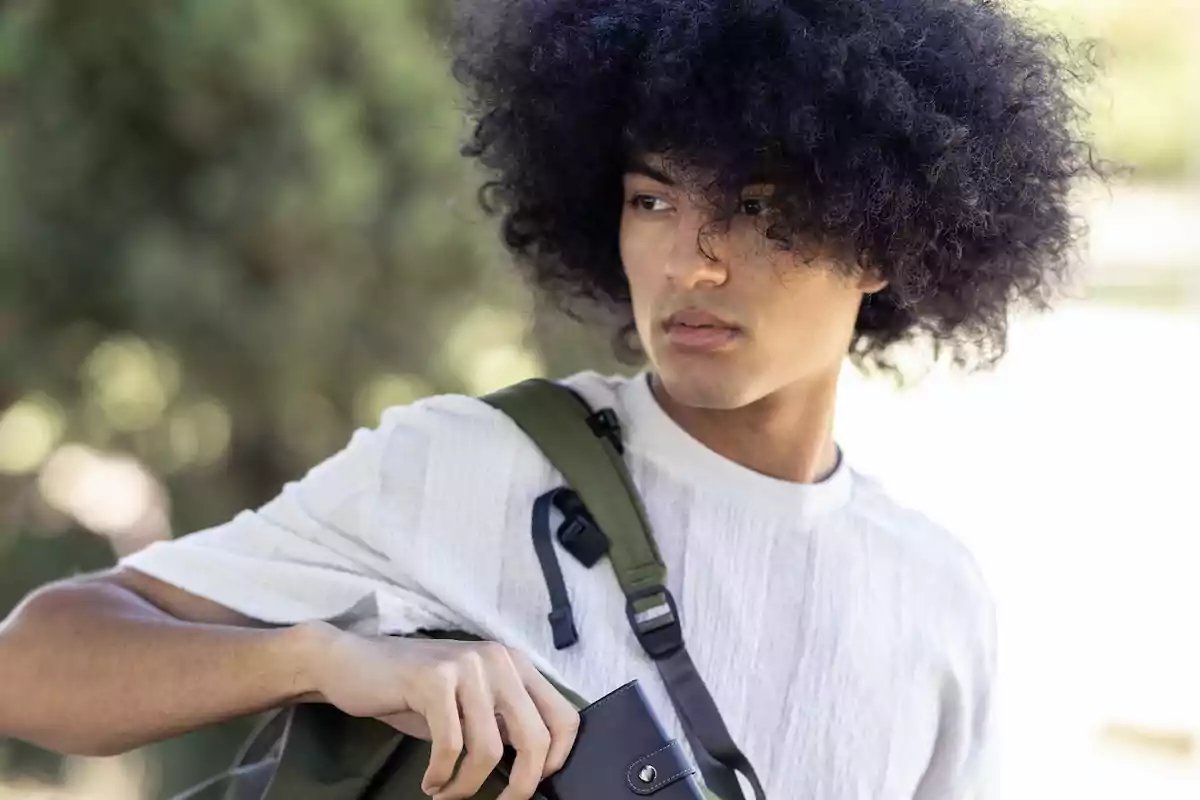 Persona con cabello rizado y voluminoso, vistiendo una camiseta blanca y llevando una mochila verde en el hombro, mirando hacia un lado en un entorno al aire libre.