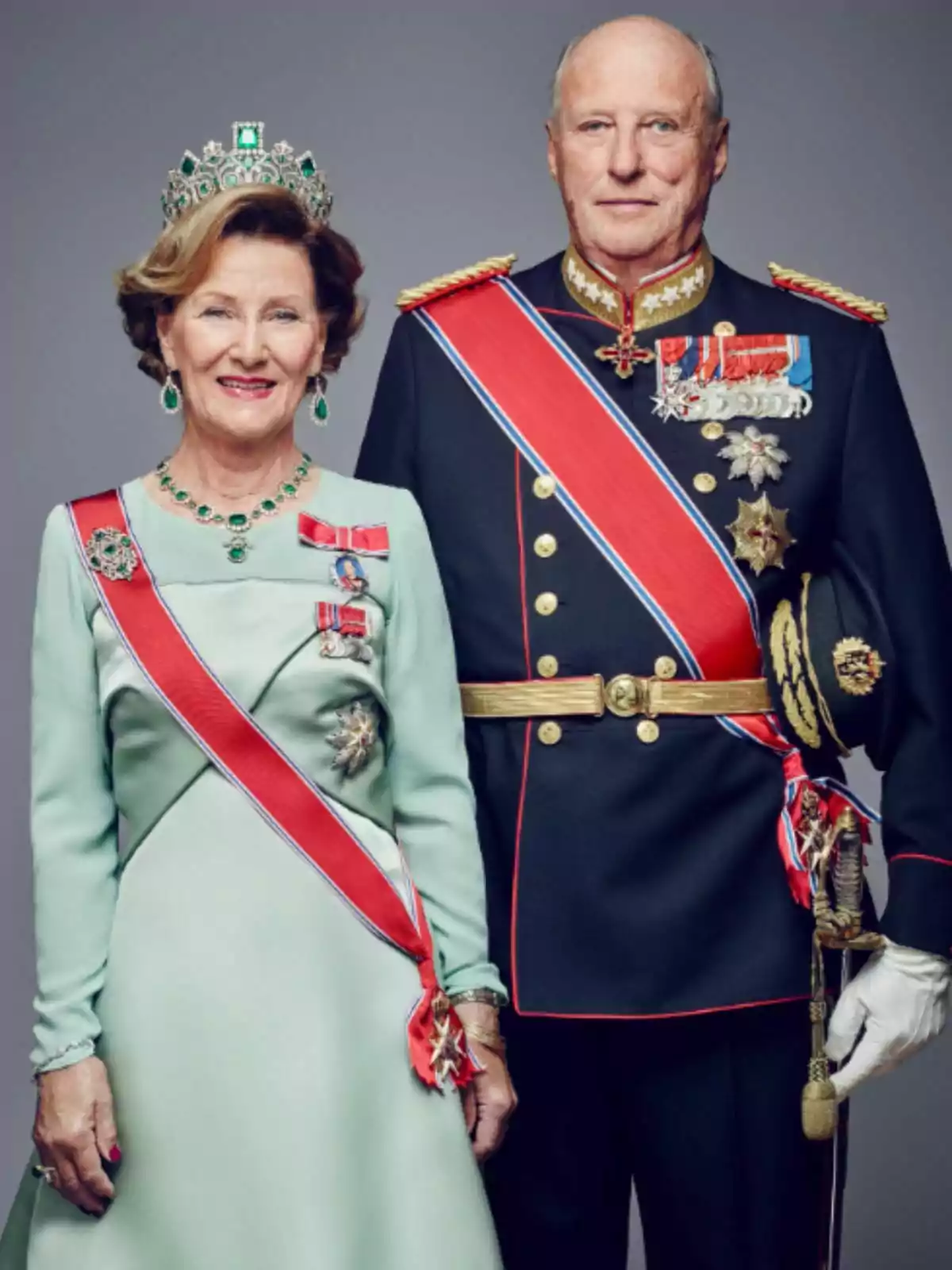 La reina Sonia y el rey Harald de Noruega vestida con trajes formales y condecoraciones, la mujer lleva una tiara y un vestido verde claro, mientras que el hombre viste un uniforme militar oscuro con medallas y una banda roja. La reina Sonia y el rey Harald de Noruega vestida con trajes formales y condecoraciones, la mujer lleva una tiara y un vestido verde claro, mientras que el hombre viste un uniforme militar oscuro con medallas y una banda roja.