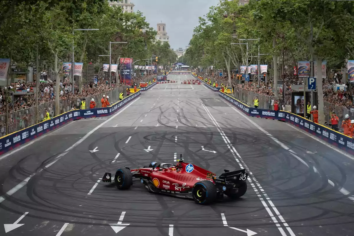 Un coche de Fórmula 1 realiza una exhibición en una calle de la ciudad, rodeado de espectadores y personal de seguridad. La calle está marcada con señales de tráfico y rodeada de árboles y edificios.