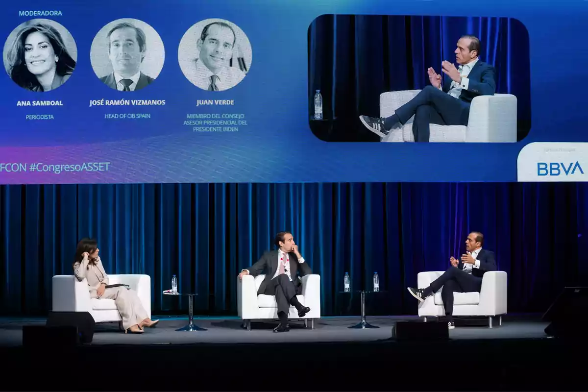 Tres personas están sentadas en un escenario participando en un panel de discusión, con una pantalla grande detrás que muestra imágenes y nombres de los participantes.