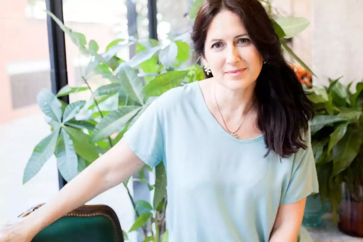 Una mujer con cabello oscuro y una blusa azul claro está sentada frente a una ventana con plantas verdes al fondo.