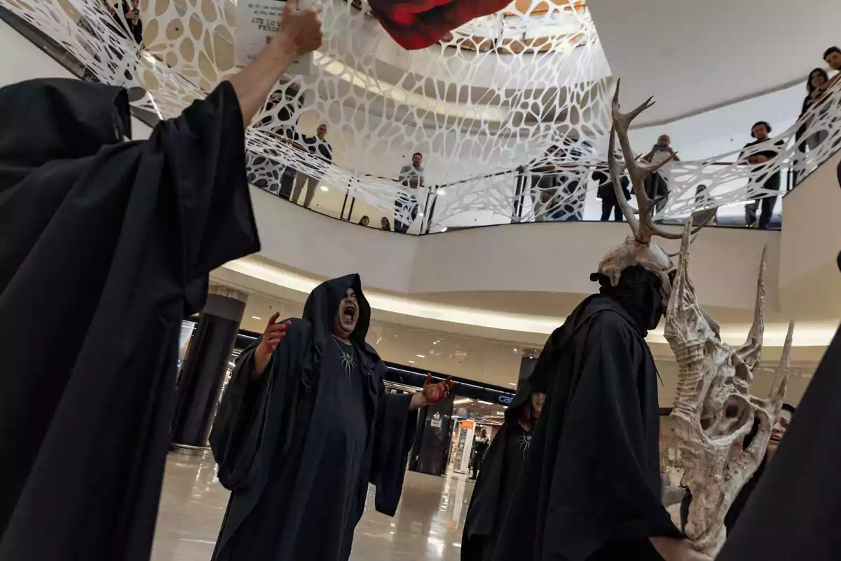 Personas con túnicas negras realizan una performance en un centro comercial, mientras el público observa desde el piso superior.