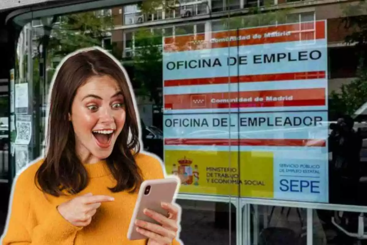 Una mujer sonriente con un suéter amarillo sostiene un teléfono móvil frente a una oficina de empleo en Madrid.