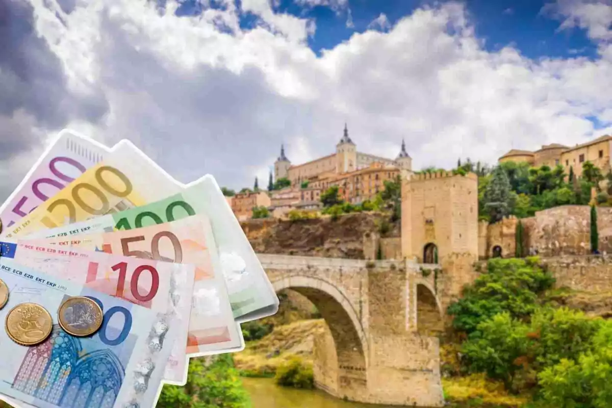 Billetes y monedas de euro frente a un puente y una ciudad histórica en un día nublado.