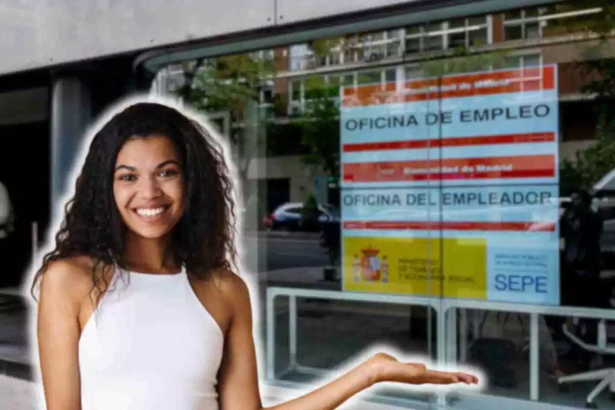 Una mujer sonriente con una camiseta sin mangas blanca está de pie frente a una oficina de empleo en Madrid, España. La oficina tiene un cartel en la ventana que dice "OFICINA DE EMPLEO" y "OFICINA DEL EMPLEADOR", junto con logotipos del Ministerio de Trabajo y Economía Social y del SEPE (Servicio Público de Empleo Estatal). La mujer parece estar señalando hacia la oficina con una mano extendida.