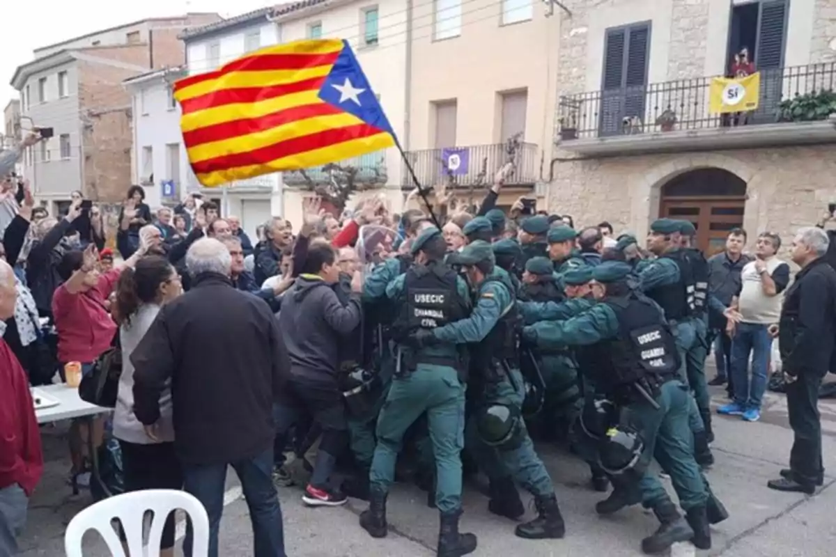 Un grupo de agentes de la Guardia Civil empuja a una multitud de personas que sostiene una bandera estelada en una calle de un pueblo, mientras varios observadores miran la escena. Un grupo de agentes de la Guardia Civil empuja a una multitud de personas que sostiene una bandera estelada en una calle de un pueblo, mientras varios observadores miran la escena.