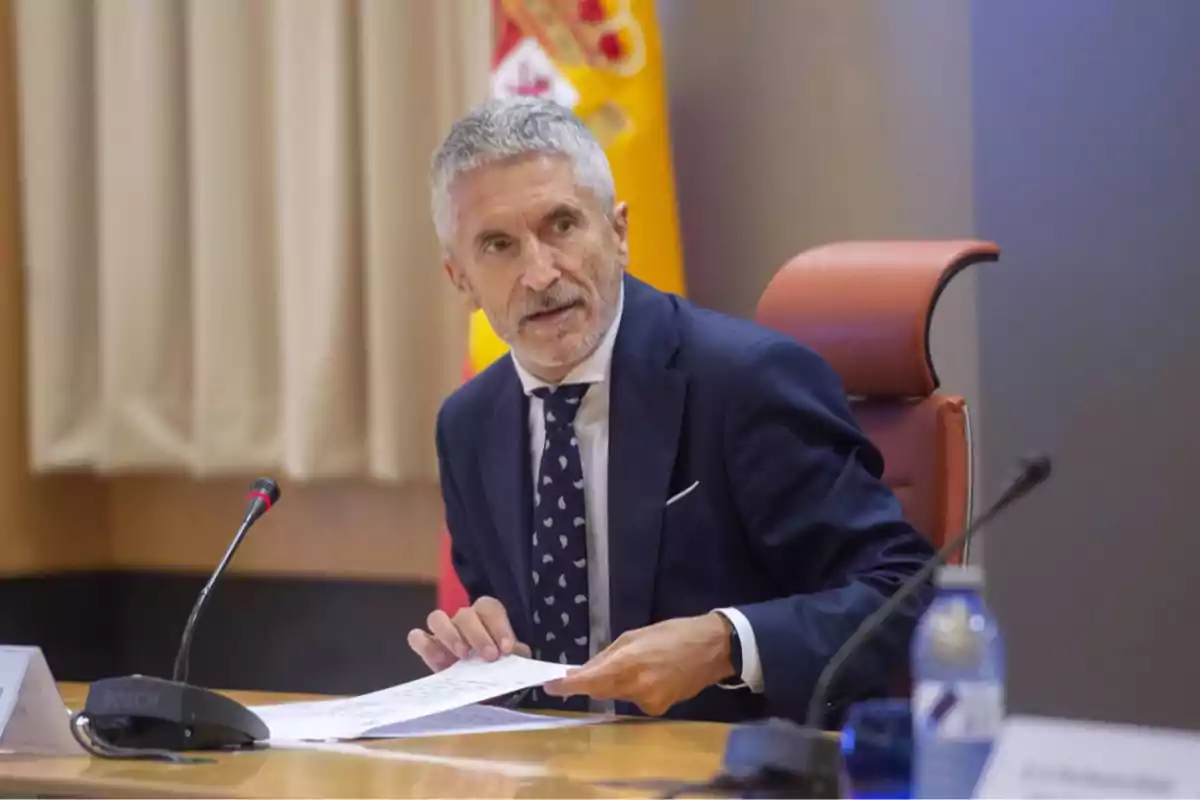 Un hombre de traje oscuro y corbata con patrones está sentado en una mesa con un micrófono y papeles en la mano, frente a una bandera. Un hombre de traje oscuro y corbata con patrones está sentado en una mesa con un micrófono y papeles en la mano, frente a una bandera.