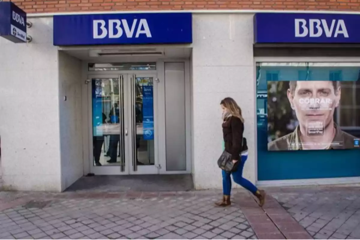 Una persona caminando frente a la entrada de una sucursal del banco BBVA.