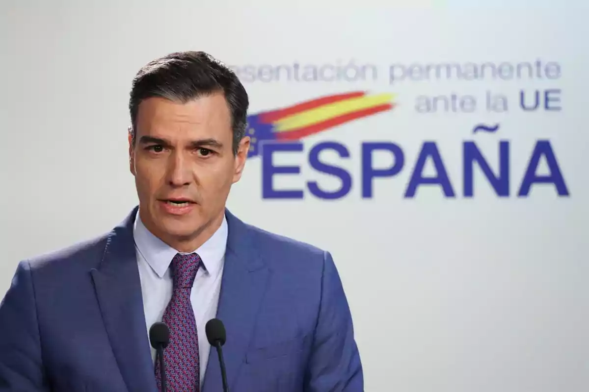 Un hombre con traje azul y corbata roja hablando frente a un micrófono con un cartel de fondo que dice "Representación permanente ante la UE ESPAÑA".