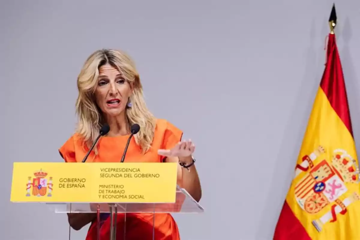 Una persona hablando en un podio con un micrófono y una bandera de España al fondo. Una persona hablando en un podio con un micrófono y una bandera de España al fondo.