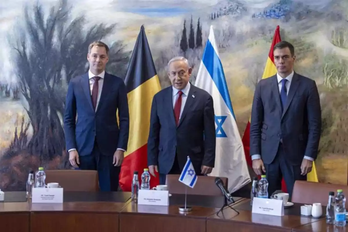 Tres hombres de traje posan frente a banderas de Bélgica, Israel y España en una sala de conferencias con un fondo de paisaje pintado. Tres hombres de traje posan frente a banderas de Bélgica, Israel y España en una sala de conferencias con un fondo de paisaje pintado.