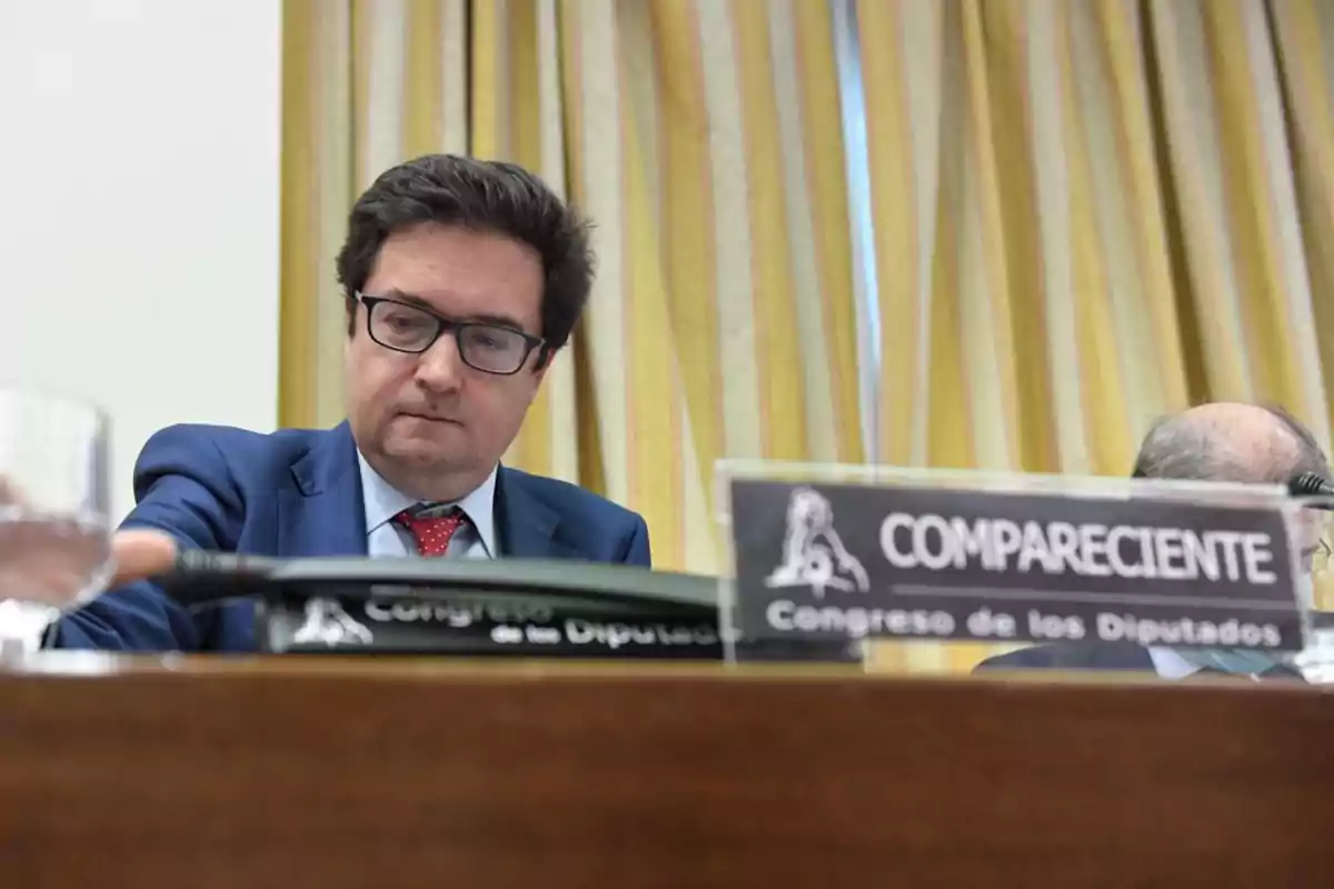 Un hombre con gafas y traje azul está sentado en una mesa con un cartel que dice Un hombre con gafas y traje azul está sentado en una mesa con un cartel que dice