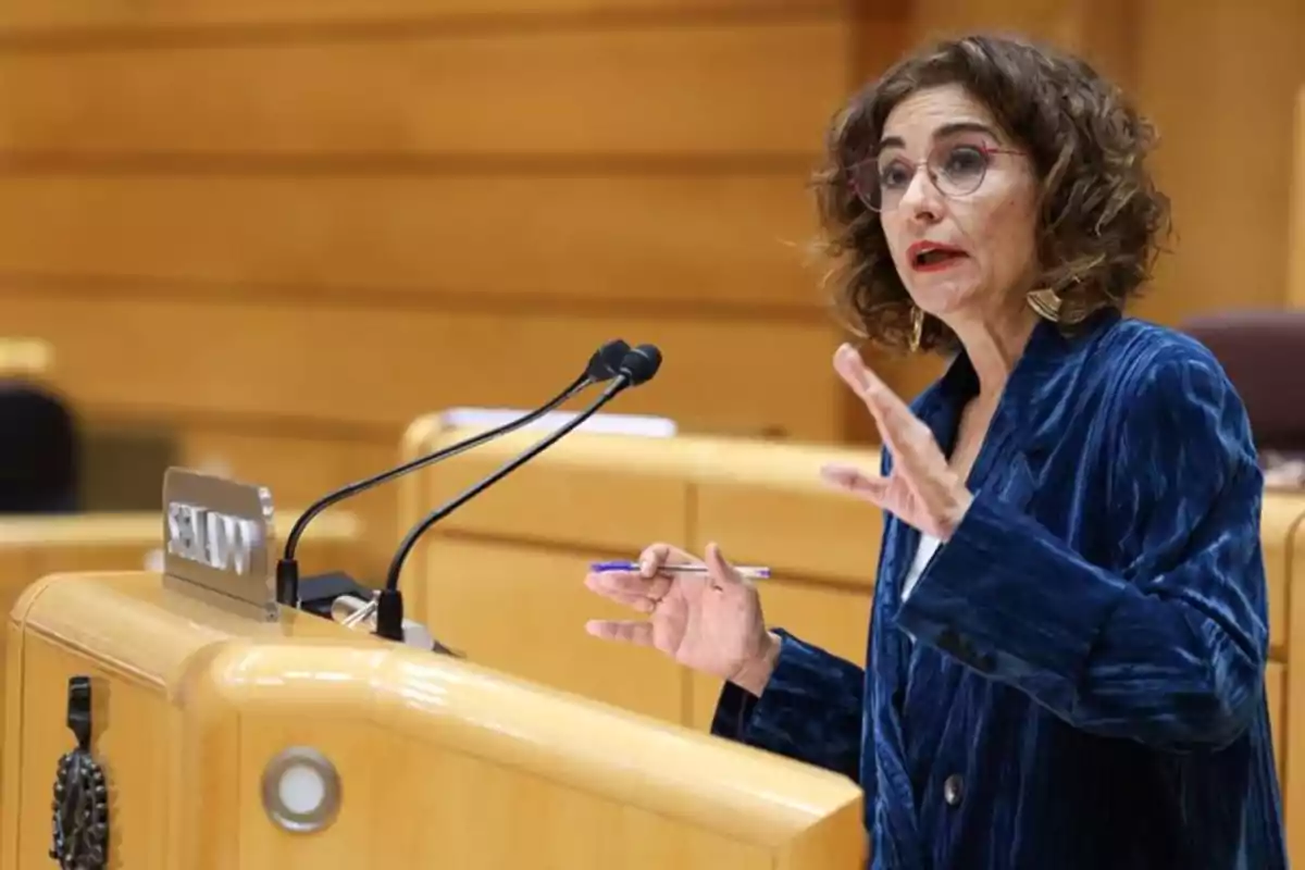 Mujer de cabello rizado y gafas hablando en un podio con micrófonos en un entorno institucional Mujer de cabello rizado y gafas hablando en un podio con micrófonos en un entorno institucional