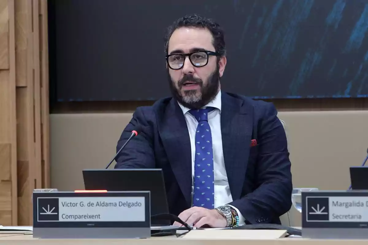 Hombre con barba y gafas hablando en una conferencia con un micrófono frente a él.