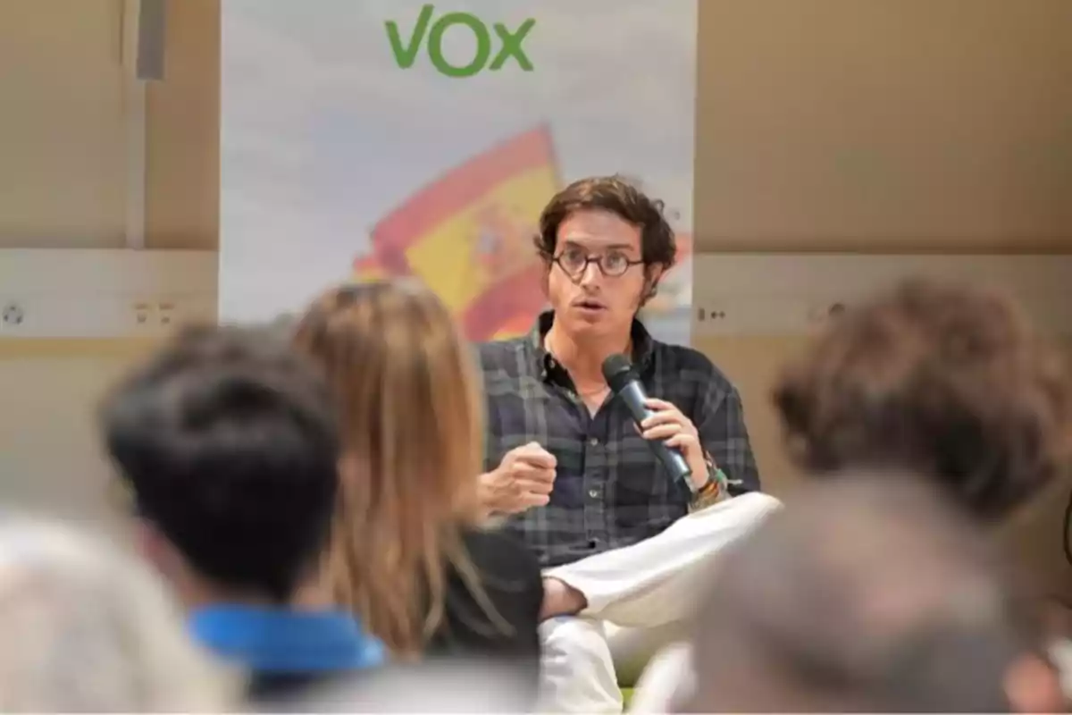 Un hombre con gafas sostiene un micrófono mientras habla frente a un grupo de personas con un cartel de Vox y una bandera de España al fondo Un hombre con gafas sostiene un micrófono mientras habla frente a un grupo de personas con un cartel de Vox y una bandera de España al fondo