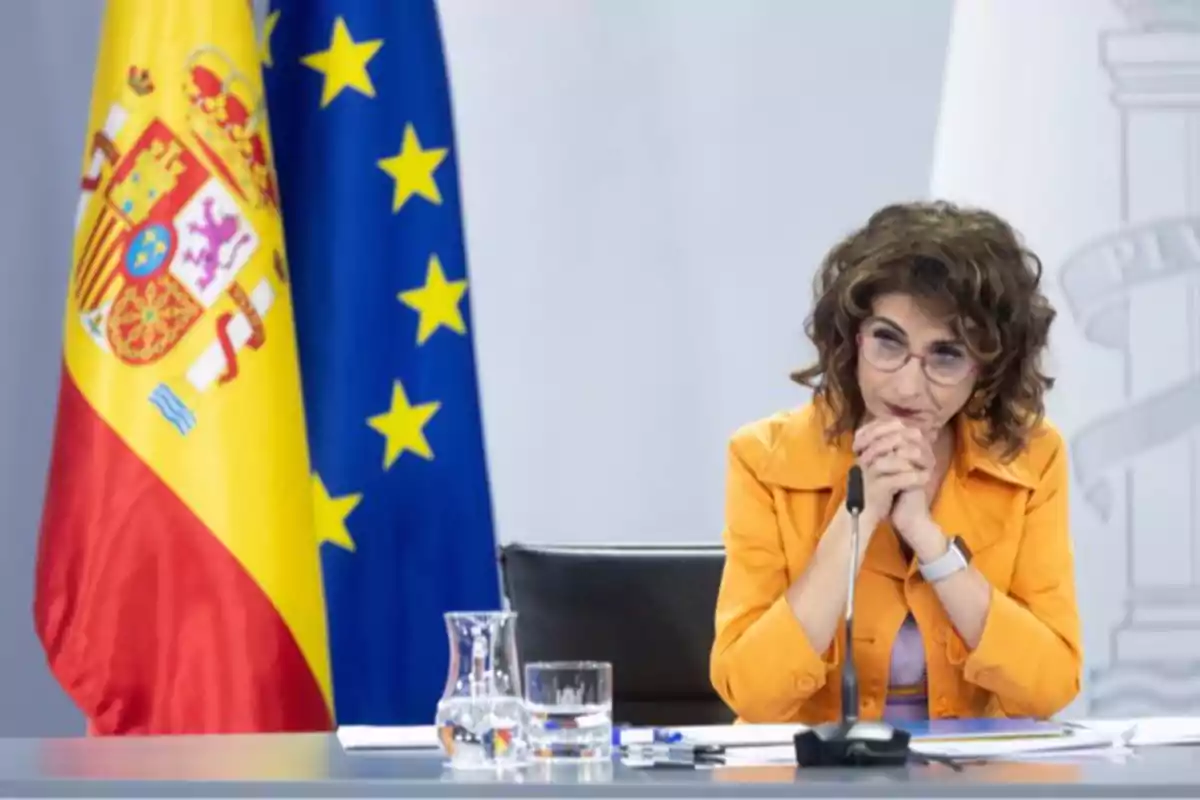 Mujer con gafas y chaqueta naranja sentada en una mesa frente a micrófono con banderas de España y la Unión Europea al fondo Mujer con gafas y chaqueta naranja sentada en una mesa frente a micrófono con banderas de España y la Unión Europea al fondo