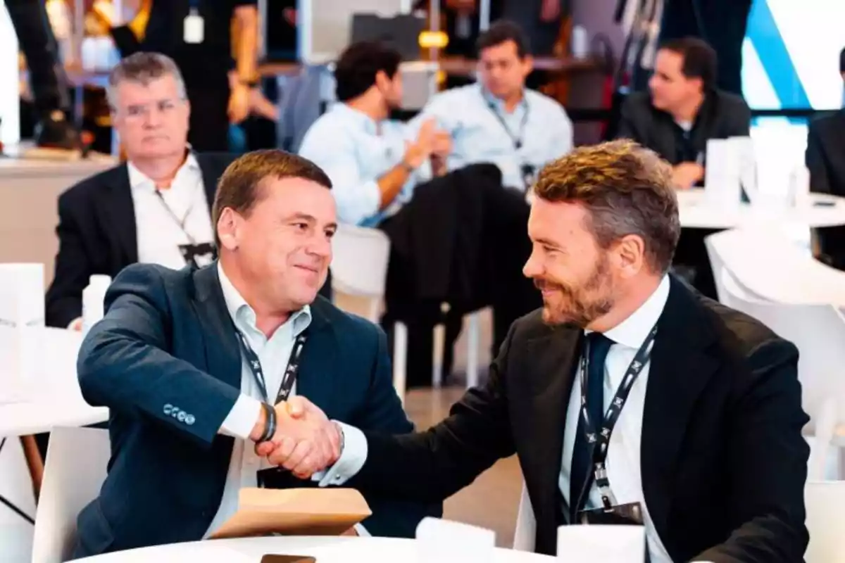 Dos hombres de negocios se dan la mano en un evento mientras otros asistentes conversan en el fondo.