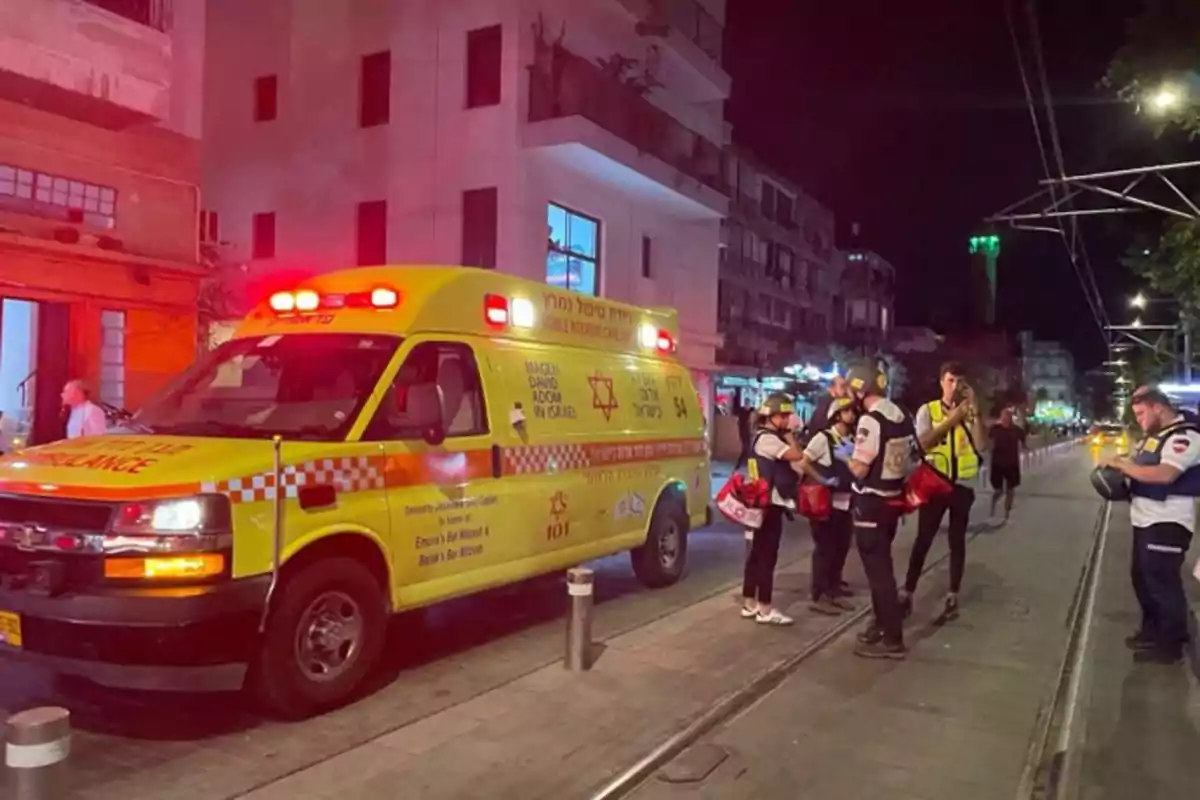 Ambulancia amarilla con luces rojas encendidas estacionada en una calle de noche con varios paramédicos alrededor.