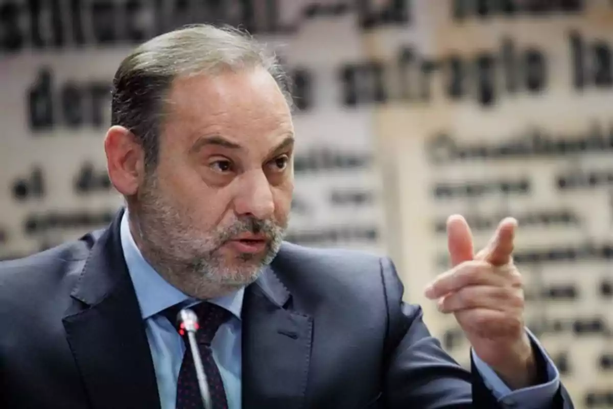Un hombre de traje y corbata gesticula mientras habla en un entorno con fondo borroso de texto. Un hombre de traje y corbata gesticula mientras habla en un entorno con fondo borroso de texto.