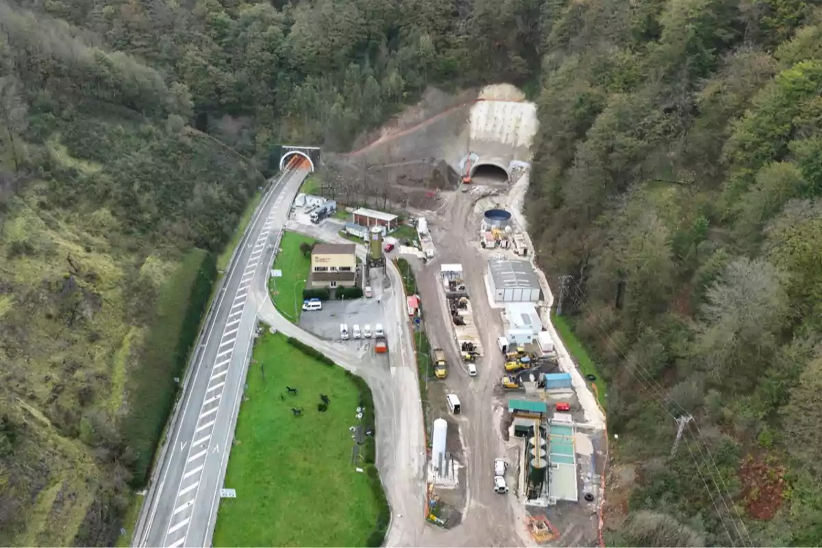 Vista aérea de una carretera con un túnel en construcción rodeado de vegetación y maquinaria de obra. Vista aérea de una carretera con un túnel en construcción rodeado de vegetación y maquinaria de obra.