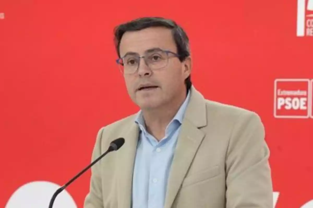 Hombre con gafas y chaqueta beige hablando frente a un micrófono con fondo rojo y logotipos del PSOE Hombre con gafas y chaqueta beige hablando frente a un micrófono con fondo rojo y logotipos del PSOE
