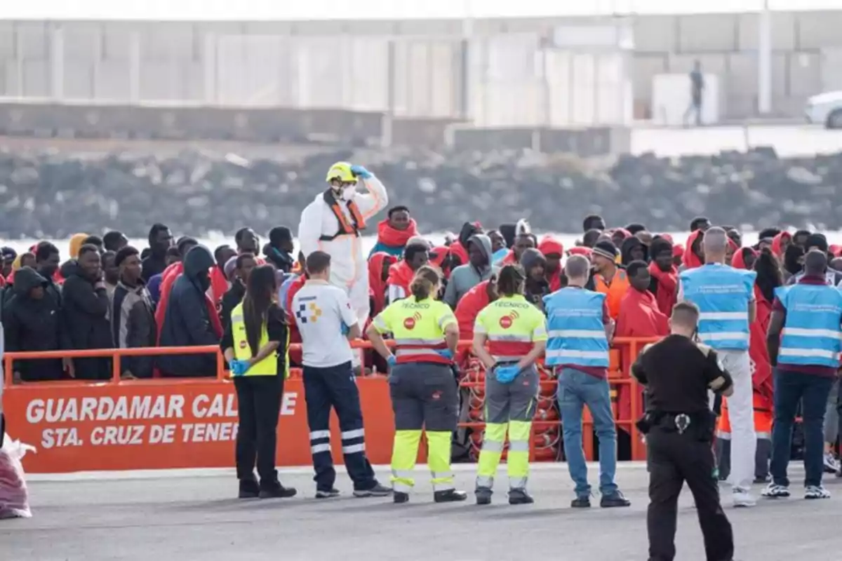 Un grupo de personas, algunas con chalecos de seguridad y otras con mantas rojas, se encuentran en un muelle junto a un cartel que dice Un grupo de personas, algunas con chalecos de seguridad y otras con mantas rojas, se encuentran en un muelle junto a un cartel que dice