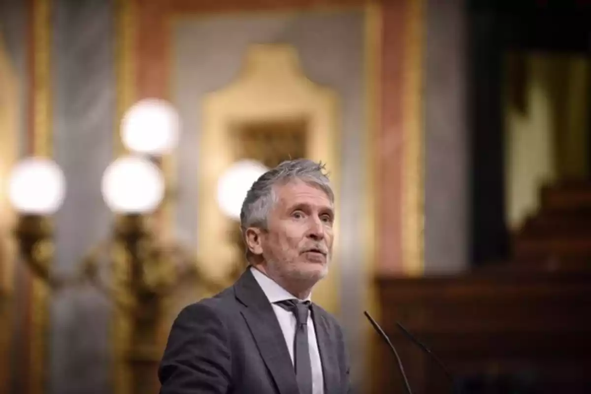 Un hombre de cabello canoso y barba está hablando en un podio en un entorno formal con luces y decoración elegante al fondo. Un hombre de cabello canoso y barba está hablando en un podio en un entorno formal con luces y decoración elegante al fondo.