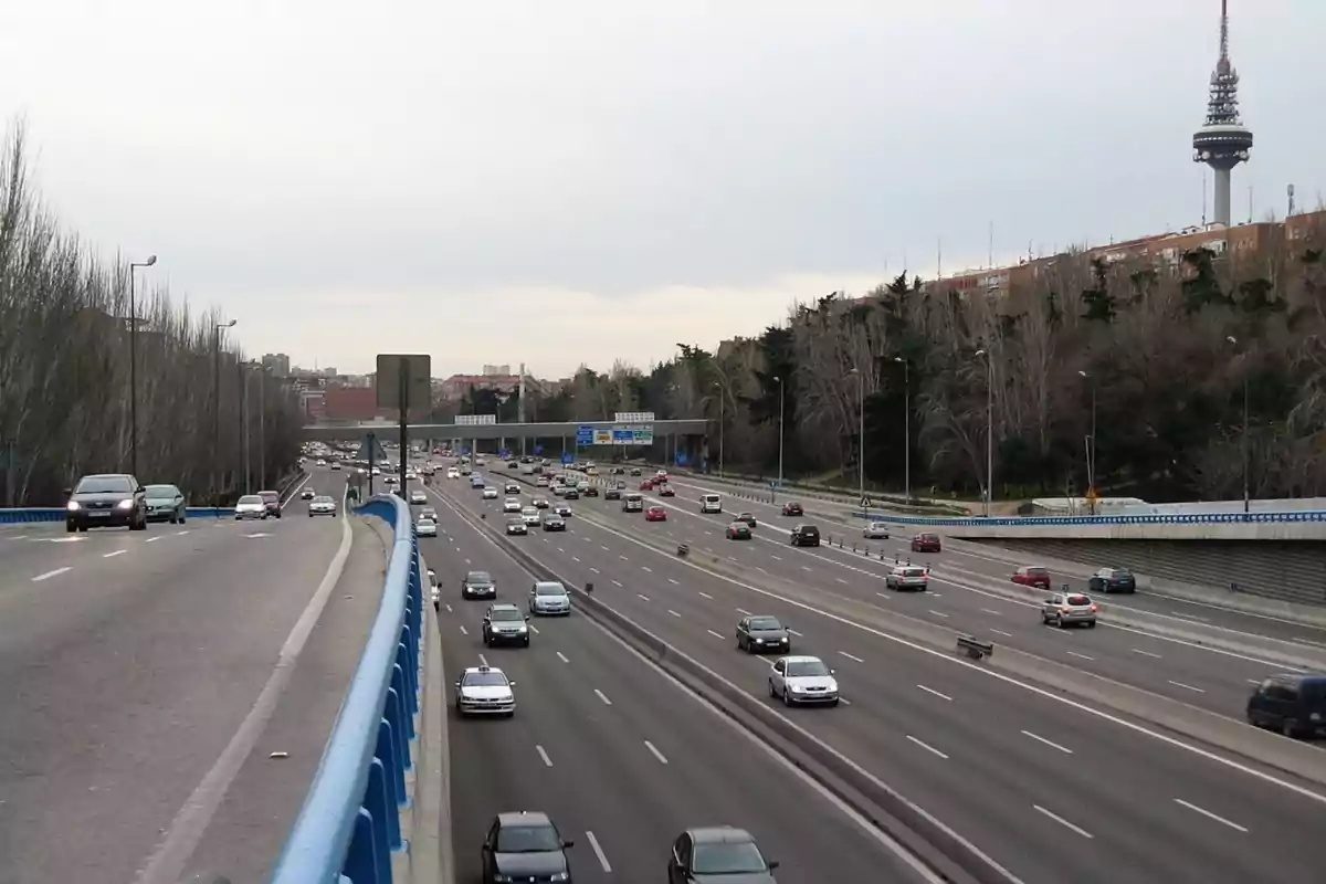 Autopista con varios carriles y tráfico moderado, con una torre de telecomunicaciones visible al fondo y árboles a los lados.
