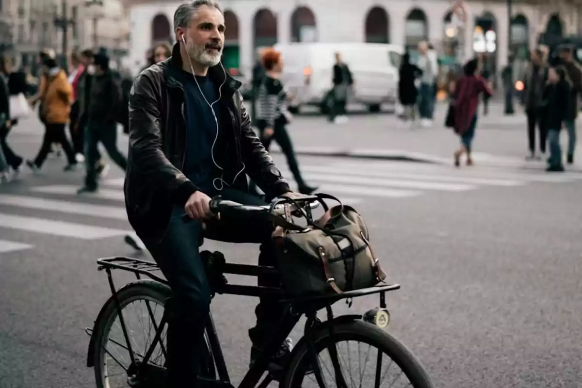 Un hombre con barba gris y auriculares monta una bicicleta en una calle concurrida, llevando una bolsa en la parte delantera.