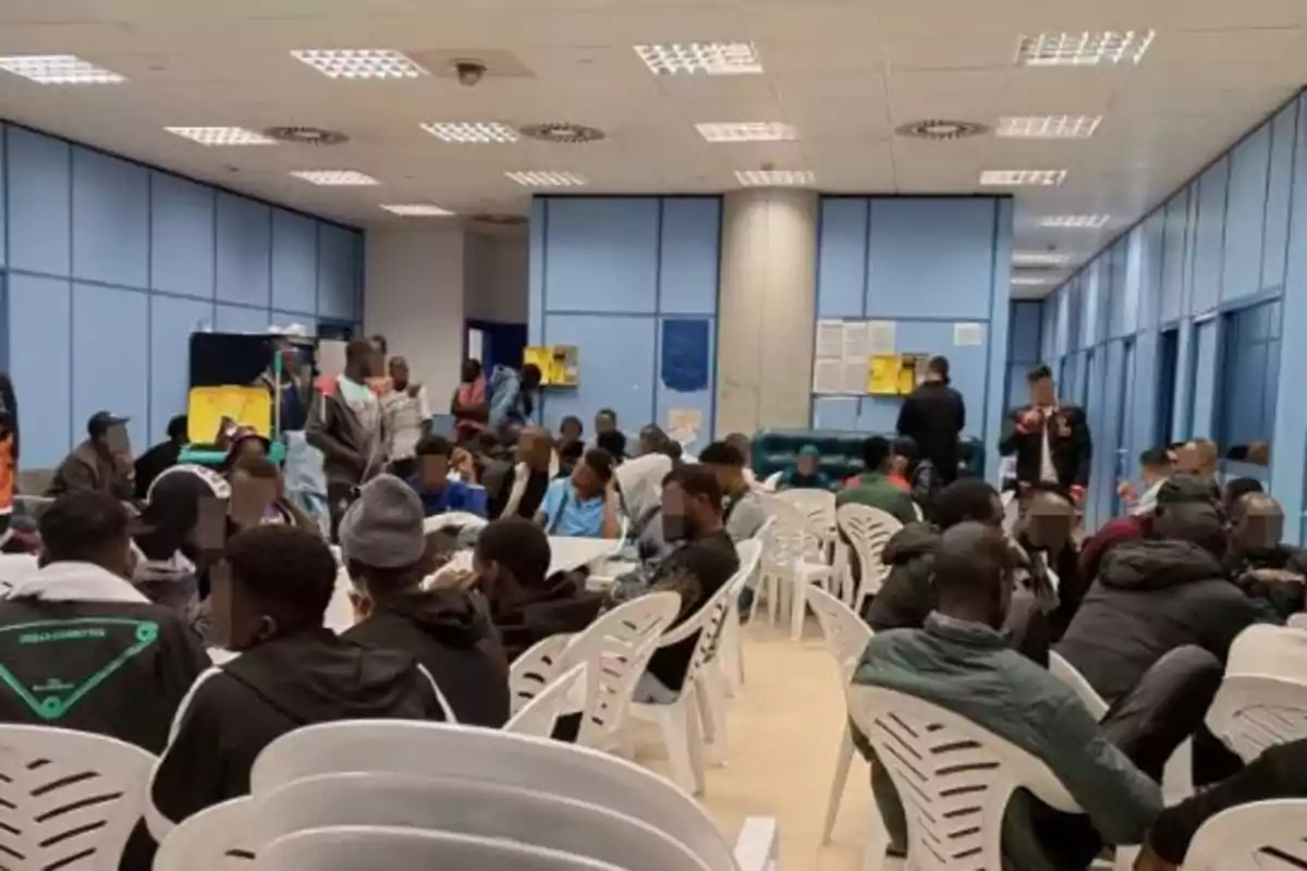 Un grupo de personas sentadas en sillas de plástico en una sala de espera con paredes azules.