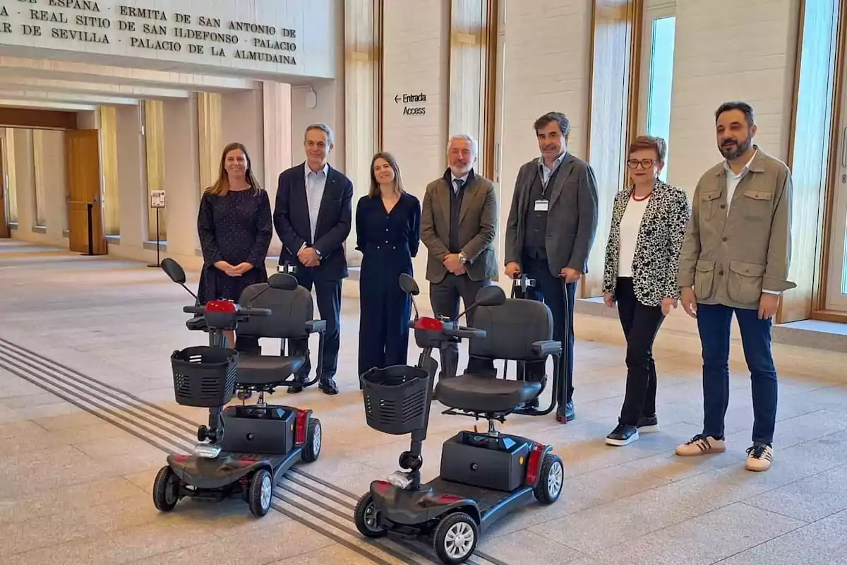 A group of people poses next to mobility scooters in a building with large windows and a sign on the wall.