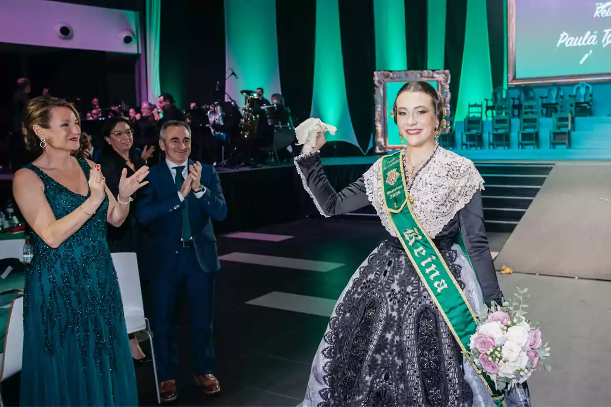 A woman dressed in a traditional outfit with a sash that says "Queen" holds a bouquet of flowers while being applauded by people at an event with green lighting and an orchestra in the background.