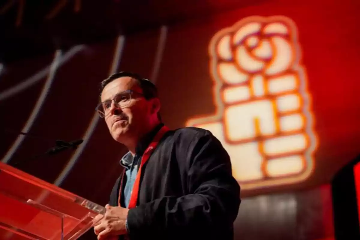 Hombre con gafas hablando en un atril con fondo rojo y un logotipo de puño y rosa iluminado detrás Hombre con gafas hablando en un atril con fondo rojo y un logotipo de puño y rosa iluminado detrás
