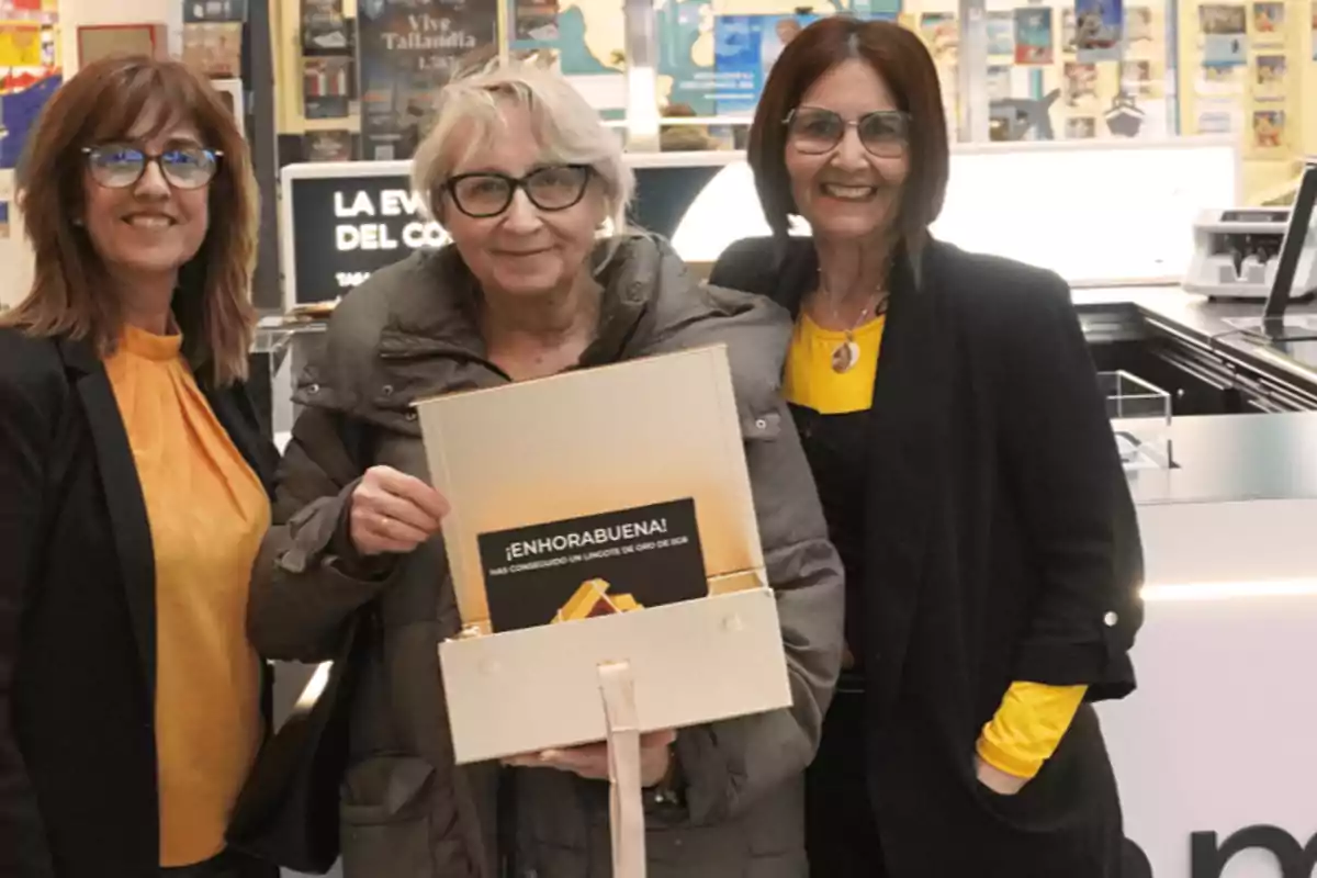 Tres mujeres sonríen mientras una de ellas sostiene una caja con un mensaje de felicitación en un entorno comercial.