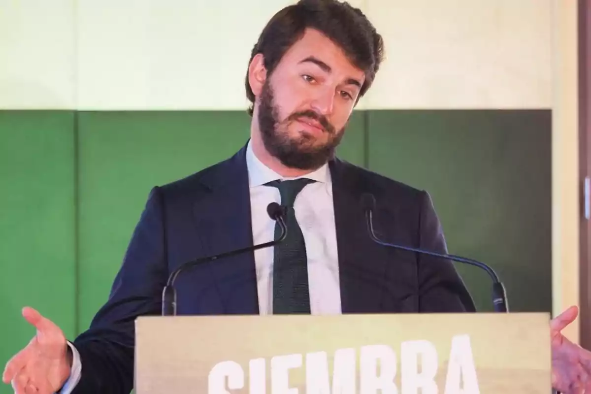 A man with a beard and a dark suit is speaking at a podium with the word "SIEMBRA" visible while gesturing with his hands.