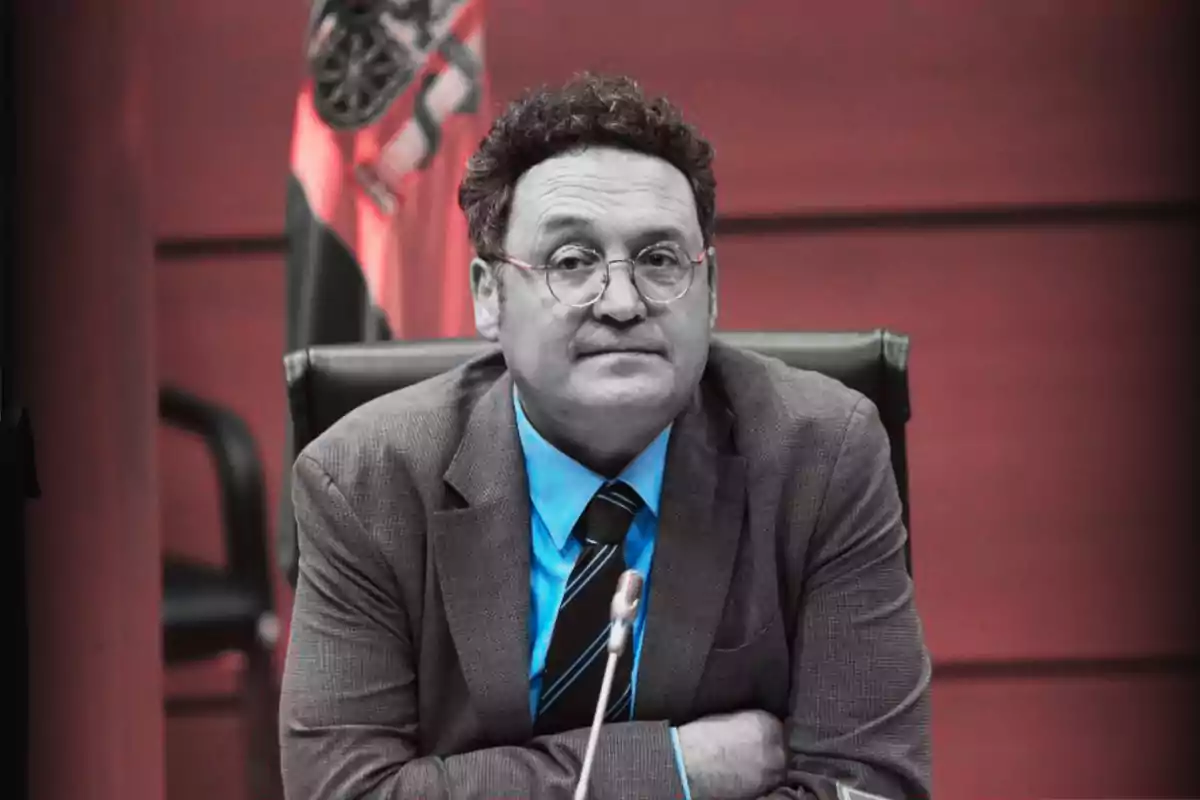 Hombre de cabello rizado y gafas sentado en una silla frente a un micrófono con fondo rojo y una bandera detrás