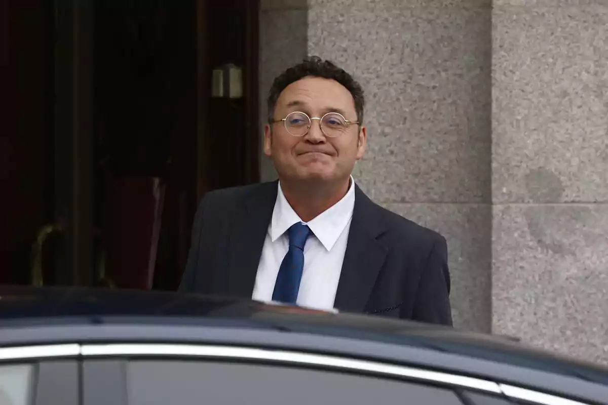 A man in glasses and a suit smiles as he stands next to a car in front of a stone building.