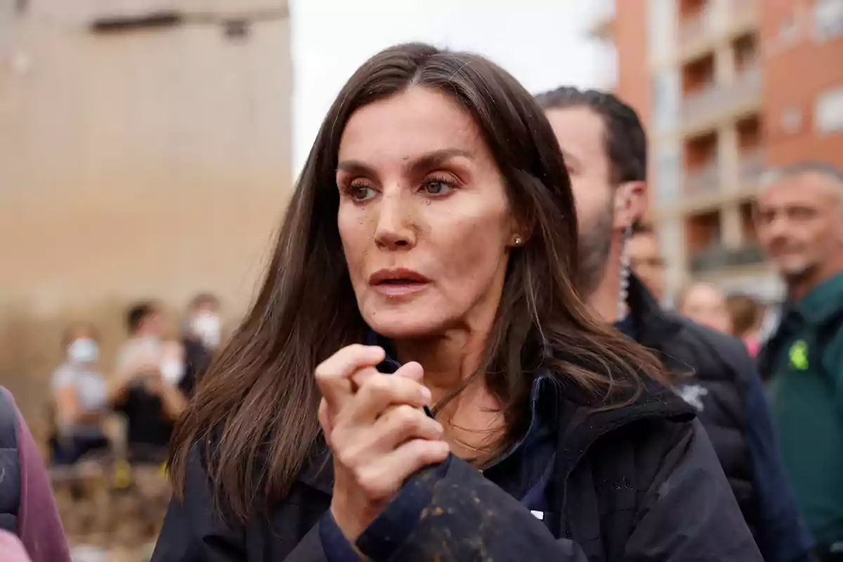 Mujer de cabello oscuro con expresión seria rodeada de personas en un entorno urbano.